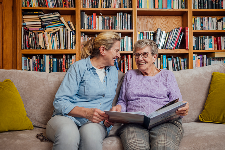 An older adult female holding a book and sitting on a coach with her caregiver in a library.