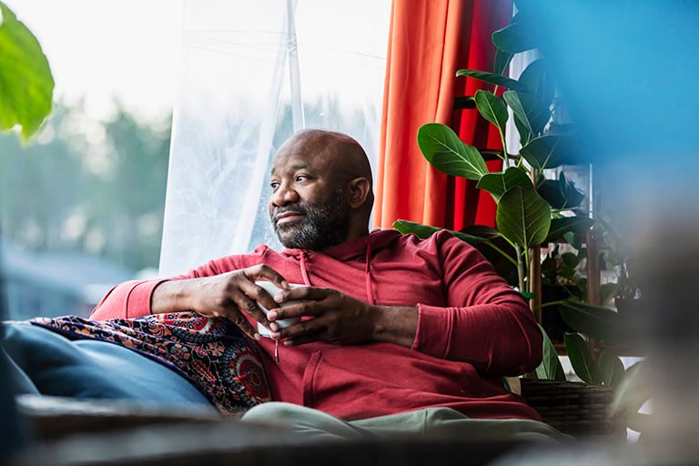 An older man at home, sitting on a couch near a window, holding a cup of tea. He is leaning back, looking away, thinking, with a serious but relaxed expression.